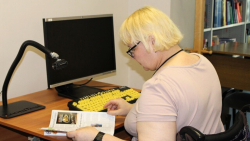 SIVA bibliotēkas lasītāja ratiņkrēslā šķirsta žurnālu pie pielāgota datorgalda