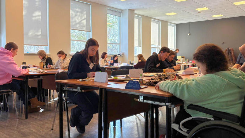 2. Cilvēki sēž pie galda un strādā ar rokdarbiem. Telpā ir lieli logi un daudz gaismas.