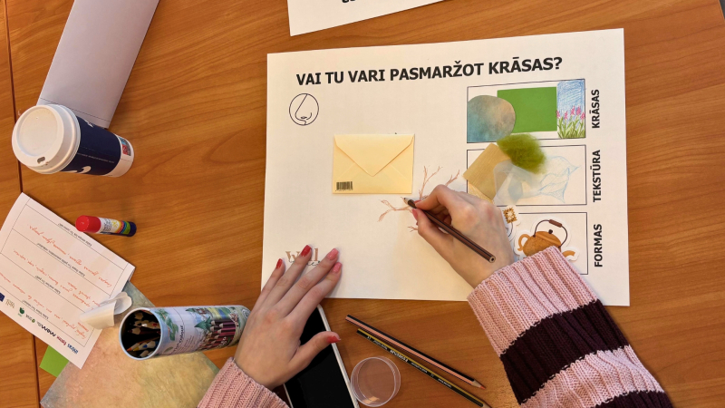 3. Cilvēks pie galda zīmē uz lapas. Uz galda ir krāsas, papīrs un citi radoši materiāli.