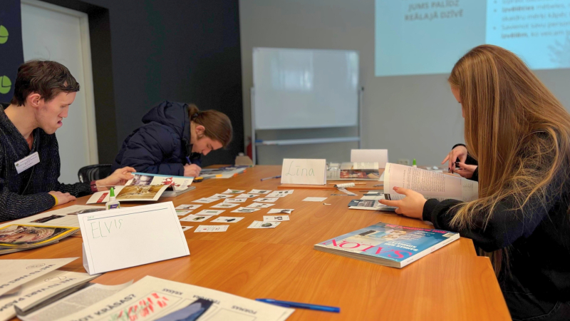 4. Trīs cilvēki sēž pie galda un strādā ar papīru un attēliem. Uz galda ir kartītes, grāmatas un līme.